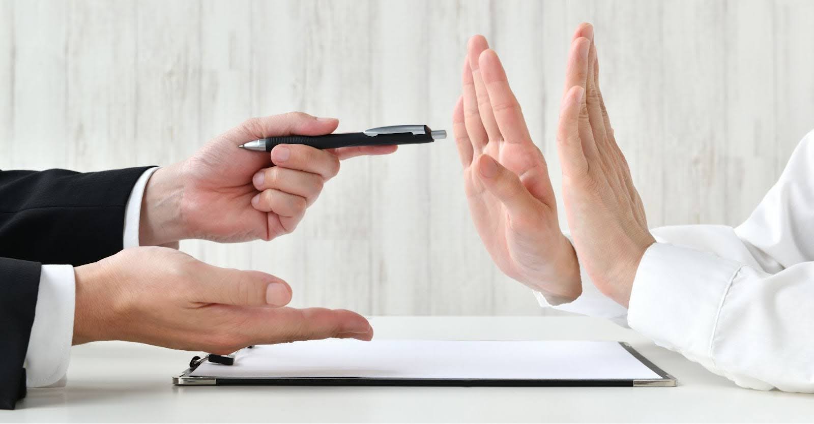 ペンを差し出しサインを迫るビジネスマンの手と、両手の平を向けて拒否する手。