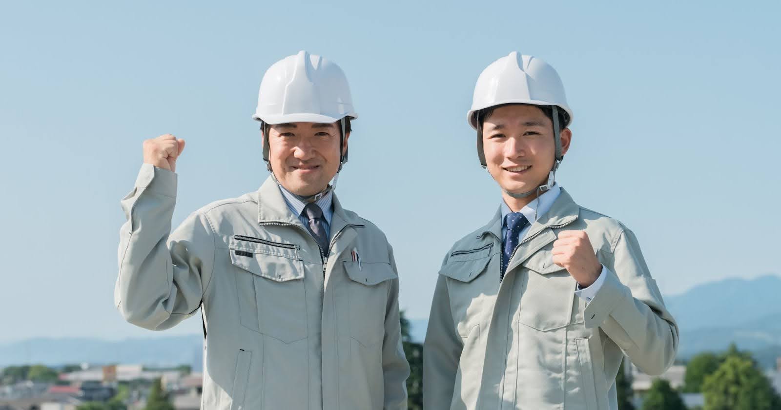 青空の下、山と街並みを背景に並んでいる作業員姿の男性二人。どちらもガッツポーズをしている。