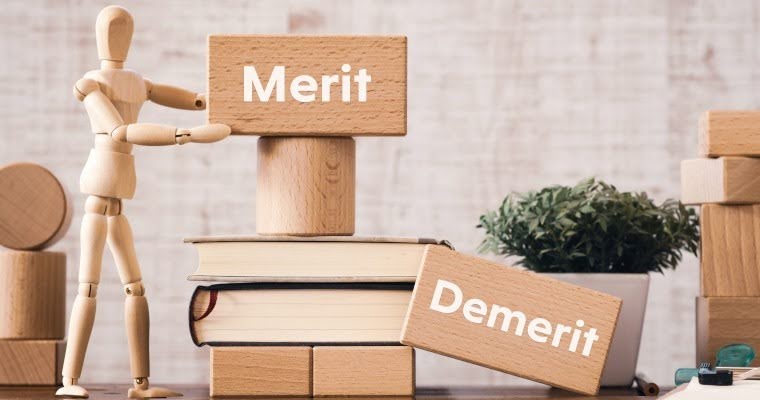 木の人形が積み木を積んでいる。「Demerit」と書かれた積み木は床に置いてあり、他の積み木や本の上に「Merit」と書かれた積み木が積まれている。