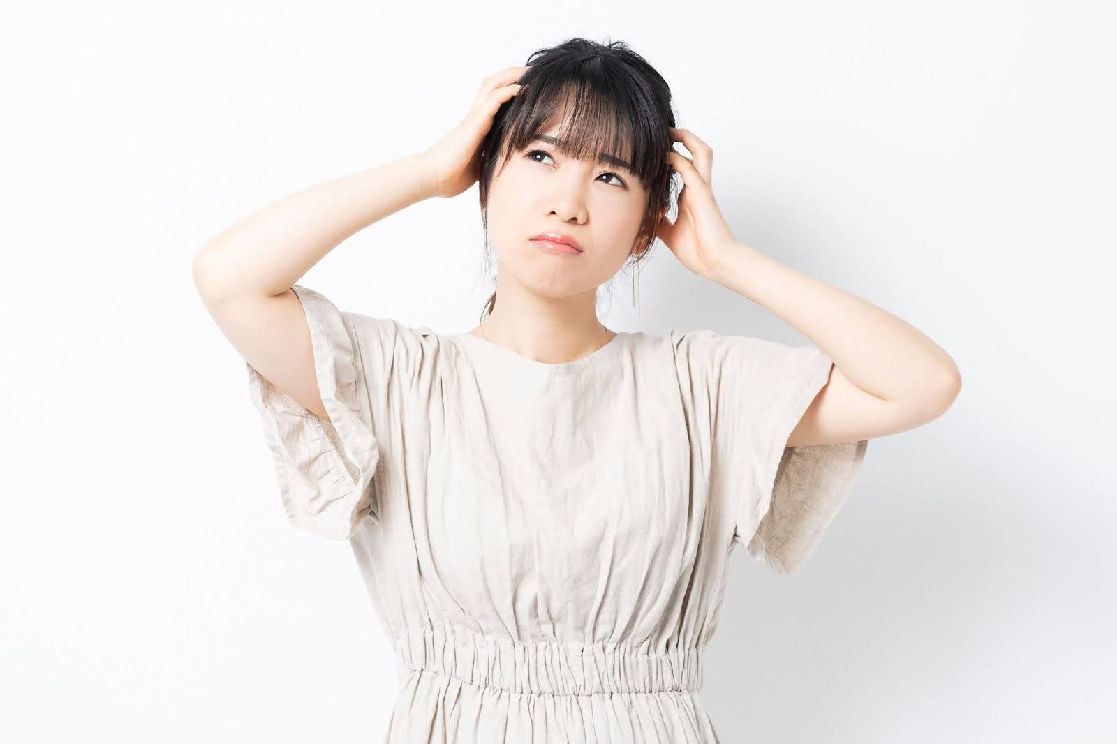 頭を両手で抱え斜め上を見つめ口をへの字に曲げているベージュの半袖ワンピースの女性