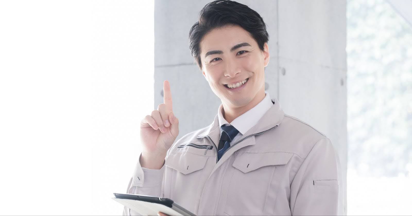 作業服姿の20代30代男性が笑顔でバインダーを持ち、こちらを見て人差し指をたてている写真