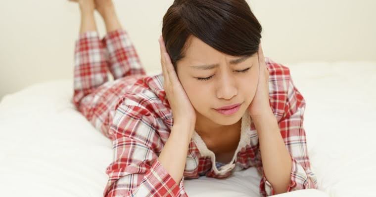 ベッドに横たわり頬杖をつきながらつらそうな顔で耳をふさぐ女性