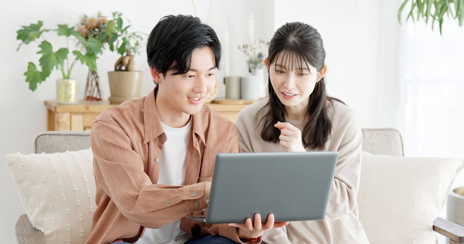 室内でノートパソコンの画面を眺め、横に座る男女。男性はノートパソコンを手で持ち、女性に画面を見せている。