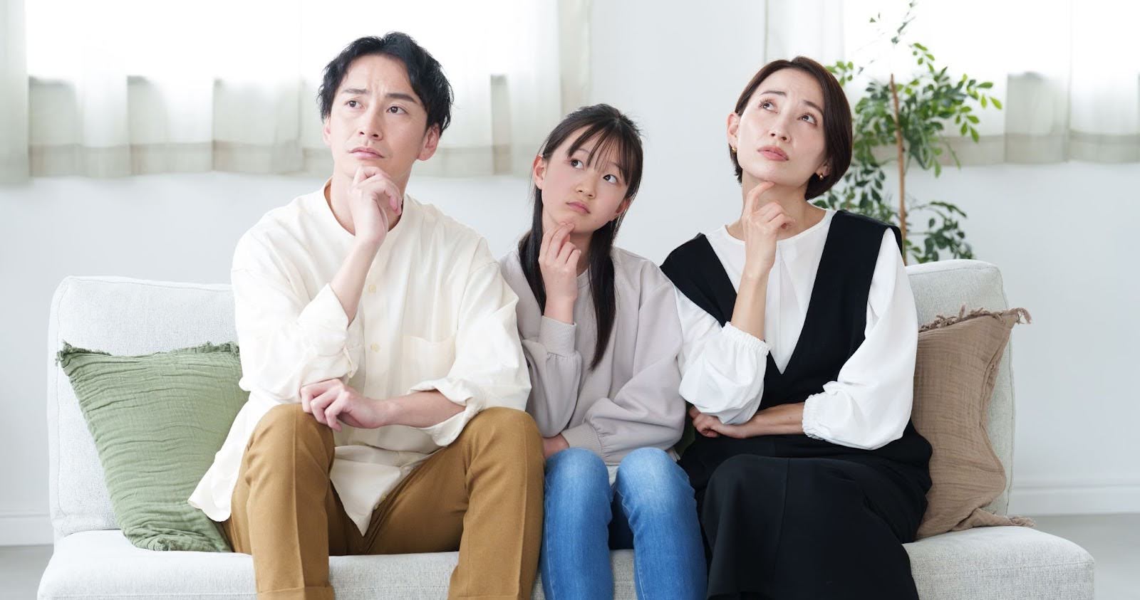 白い壁のリビング、白いソファに座る三人。左から順に、父親、女の子、母親。三人とも顎付近に軽く握ったこぶしをあて、左斜め上を見つめながら、首をかしげている。